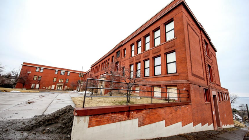 A large red brick building with numerous windows sits on a sloped sidewalk next to a grassy area and a wheelchair ramp; another similar building is visible in the background under a cloudy sky.