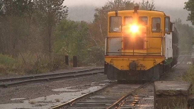 A yellow train engine with its headlight shining approaches on wet railroad tracks, surrounded by trees and greenery on a cloudy day.