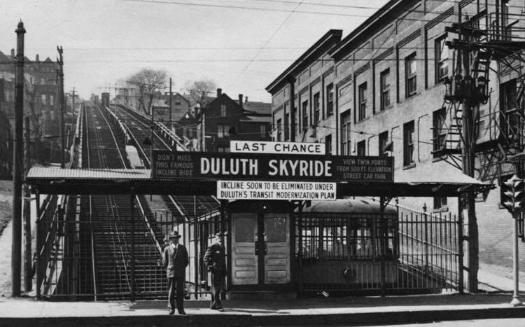 Historic black-and-white photo of the Duluth Skyride incline station, with a sign warning it will soon be eliminated. Two men stand near the entrance, and tracks lead steeply uphill between buildings.