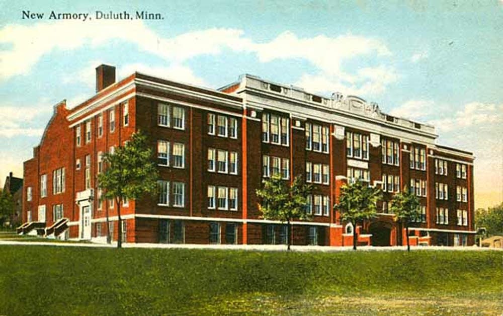 A vintage postcard shows the New Armory in Duluth, Minnesota—a large, three-story red brick building with many windows, white trim, and trees in front, set against a partly cloudy sky.