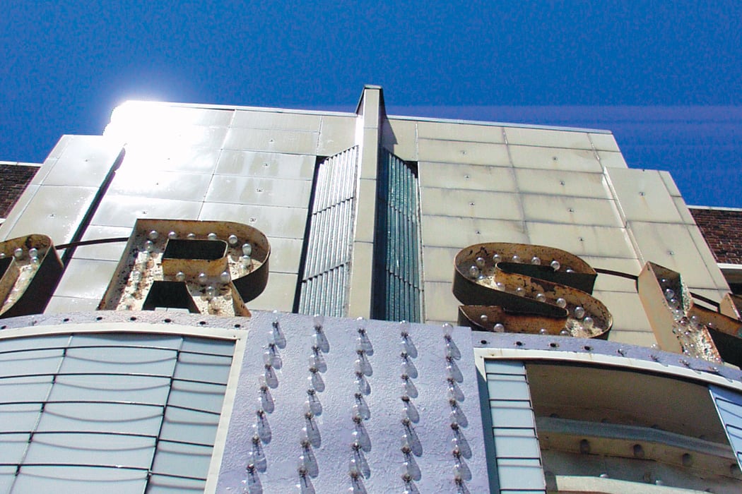 Close-up of an old, weathered theater marquee sign with rusty, partially visible letters and rows of light bulbs, set against a bright blue sky. Sunlight reflects off the metal surface.