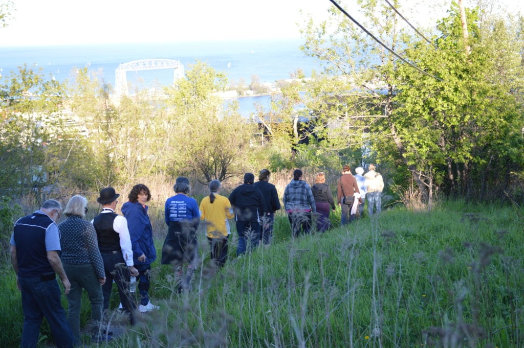 A group of people walk single file down a grassy hill surrounded by trees, with a body of water and a large bridge visible in the background under a bright sky.