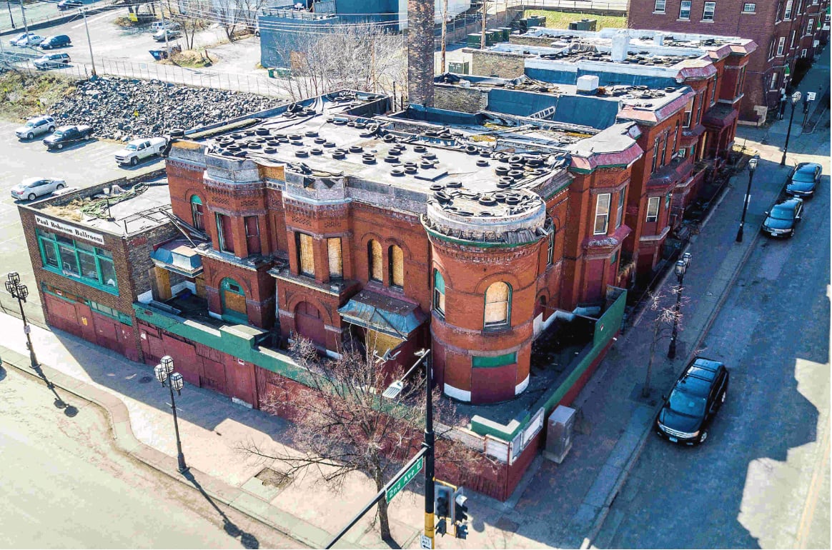 Aerial view of a large, old, red-brick building with boarded windows and damaged roof, located on a street corner with parked cars. The building appears vacant and surrounded by urban streets and empty lots.