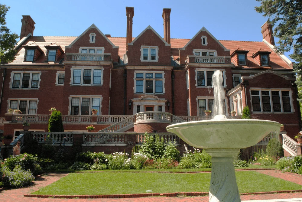 A large, historic red-brick mansion with gabled roofs and tall chimneys stands behind a marble fountain and lush, landscaped gardens on a sunny day.
