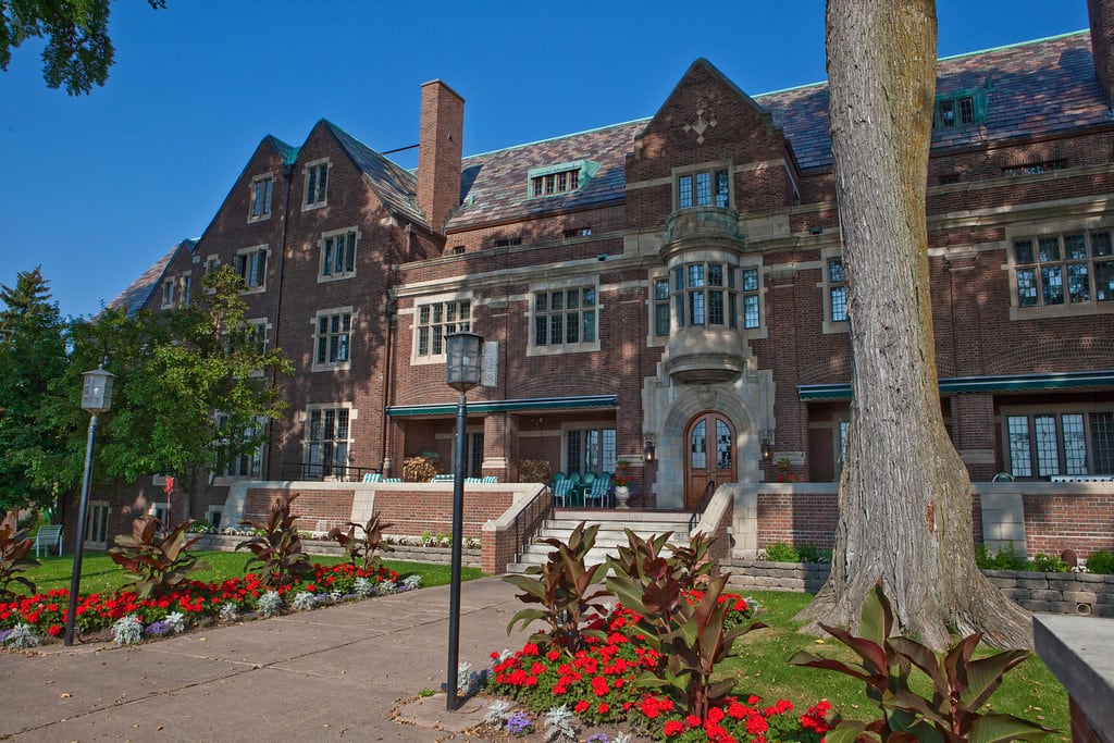 A large brick building with a steep roof and multiple windows, surrounded by well-kept gardens with red flowers, green shrubs, and large trees, under a clear blue sky.
