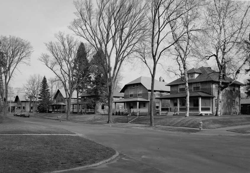 A black-and-white photo of a quiet suburban street with several large, old houses and leafless trees lining the sidewalk on a cloudy day.
