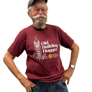 A man with a gray mustache and baseball cap wearing a maroon t-shirt that says “Old Building Hugger” with a graphic of a historic building.